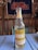 A bottle of Caribe pineapple-flavored cider sits on a blue wooden table. The bottle is clear with yellow and white labeling, featuring an apple icon. In the background, a red and white Ole Miss banner hangs on a rustic wooden wall alongside old license plates and hubcaps.
