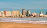 A beachfront cityscape with a line of tall buildings along the shoreline. The sandy beach in the foreground is met by gentle waves, and the sky is clear with a few clouds.