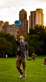 A person is skillfully spinning a blue frisbee on their finger in a park. They are surrounded by lush green grass and distant trees, with city buildings towering in the background. Others can be seen enjoying recreational activities in the park.