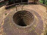A stone well with a circular opening is surrounded by moss-covered bricks. The weathered stone structure suggests an old and possibly historical setting, with a wooden pulley system above the well. The area around the well is an open courtyard with a small overhanging roof structure and a backdrop of lush greenery.