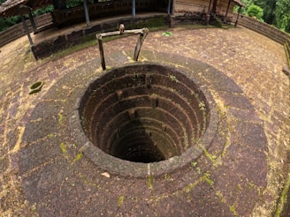 A stone well with a circular opening is surrounded by moss-covered bricks. The weathered stone structure suggests an old and possibly historical setting, with a wooden pulley system above the well. The area around the well is an open courtyard with a small overhanging roof structure and a backdrop of lush greenery.