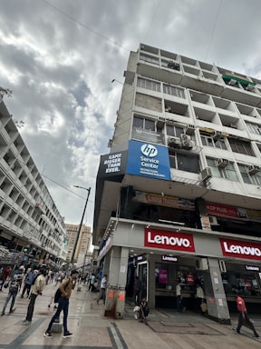 An urban street scene featuring a multi-story building with retail spaces on the ground floor, including prominent signs for Lenovo and an HP service center. The building's facade shows signs of wear, and there are several people walking along the street. The sky is filled with grey clouds, suggesting an overcast day.