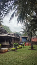 A serene outdoor setting featuring an ancient stone temple surrounded by lush greenery and tall palm trees. Several people dressed in traditional orange attire are walking near the temple structure, which is flanked by manicured shrubs and an expansive lawn. The sky is overcast, adding a tranquil ambience to the scene.