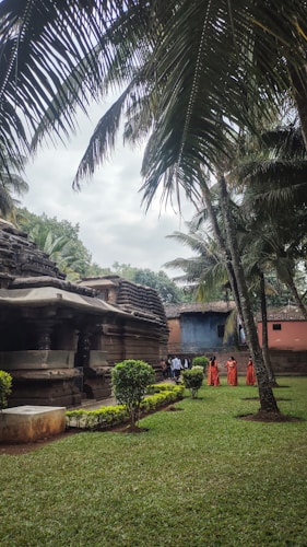 A serene outdoor setting featuring an ancient stone temple surrounded by lush greenery and tall palm trees. Several people dressed in traditional orange attire are walking near the temple structure, which is flanked by manicured shrubs and an expansive lawn. The sky is overcast, adding a tranquil ambience to the scene.
