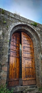 An ancient, arched wooden door set within a stone structure, partially open, revealing a glimpse of the path beyond. The door features intricate ironwork and round metal studs, while the stone wall shows signs of age with patches of moss and grass growing in the crevices. The atmosphere suggests a sense of history and mystery.