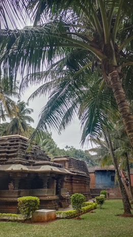 An ancient stone temple surrounded by lush green vegetation, featuring palm trees overhead. The structure has intricate carvings and is set in a serene garden with well-maintained hedges. The atmosphere appears tranquil, with a slightly overcast sky adding to the calm ambiance.