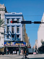 A city street scene with a prominent blue building featuring New Orleans-style architecture. The corner shop, Jazz City, offers spirits, gifts, and luggage. A cyclist rides in the foreground under a traffic light, while the street sign indicates 'Canal.' The sky is clear and blue.