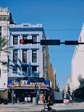 A city street scene with a prominent blue building featuring New Orleans-style architecture. The corner shop, Jazz City, offers spirits, gifts, and luggage. A cyclist rides in the foreground under a traffic light, while the street sign indicates 'Canal.' The sky is clear and blue.