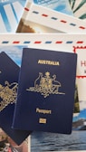 A blue Australian passport with gold embossing is placed on top of travel brochures and documents, featuring images of scenic locations and elements like blue skies and an envelope with red and blue stripes.