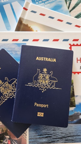 A blue Australian passport with gold embossing is placed on top of travel brochures and documents, featuring images of scenic locations and elements like blue skies and an envelope with red and blue stripes.