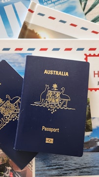 A blue Australian passport with gold embossing is placed on top of travel brochures and documents, featuring images of scenic locations and elements like blue skies and an envelope with red and blue stripes.