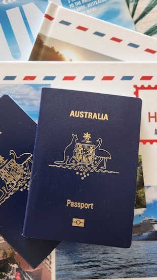 A blue Australian passport with gold embossing is placed on top of travel brochures and documents, featuring images of scenic locations and elements like blue skies and an envelope with red and blue stripes.