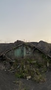 An old, abandoned house partially buried and overgrown with plants and debris. The structure shows signs of decay with its roof collapsing and greenery overtaking what remains of the walls.