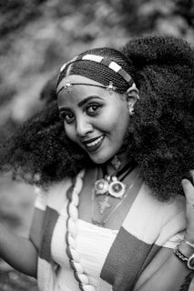 A woman smiles warmly, adorned with intricate hair braiding complemented by decorative beads. She wears traditional jewelry, including a statement necklace with large circular and cross-shaped pendants. Her attire features patterned fabric draped over her shoulders.