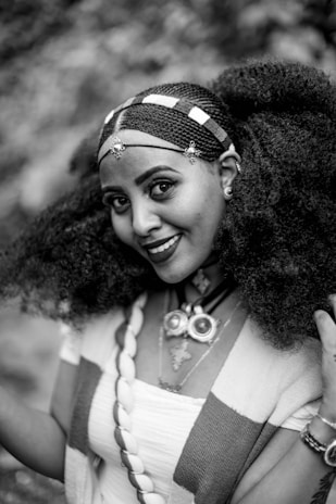 A woman smiles warmly, adorned with intricate hair braiding complemented by decorative beads. She wears traditional jewelry, including a statement necklace with large circular and cross-shaped pendants. Her attire features patterned fabric draped over her shoulders.
