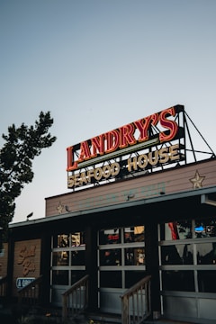 A well-lit neon sign for Landry's Seafood House is displayed on the roof of a building. The sign features bold red letters with a vintage look, and the facade below is adorned with stars. The surroundings include some trees and evening lighting, suggesting a relaxed outdoor atmosphere.