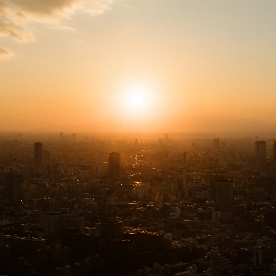 The image depicts a cityscape at sunset, with the sun setting behind the horizon and casting a warm glow over the urban landscape. The sky is painted in shades of orange and yellow, creating a stunning backdrop for the buildings that fill the scene. The perspective from above provides a bird's eye view of the city, showcasing its architectural diversity and the interplay between nature and human-made structures.