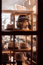 A variety of hats are displayed on wooden shelves inside a store window. A mannequin or doll dressed in traditional attire and wearing a hat is prominently featured, surrounded by several types of hats, including fedoras and bucket hats. The warm lighting creates a cozy and inviting atmosphere.