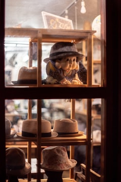 A variety of hats are displayed on wooden shelves inside a store window. A mannequin or doll dressed in traditional attire and wearing a hat is prominently featured, surrounded by several types of hats, including fedoras and bucket hats. The warm lighting creates a cozy and inviting atmosphere.