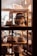 A variety of hats are displayed on wooden shelves inside a store window. A mannequin or doll dressed in traditional attire and wearing a hat is prominently featured, surrounded by several types of hats, including fedoras and bucket hats. The warm lighting creates a cozy and inviting atmosphere.