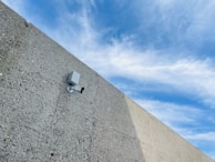 A security camera mounted on a rough, textured concrete wall, angled slightly upwards. The background shows a partly cloudy sky with various shades of blue and white clouds.
