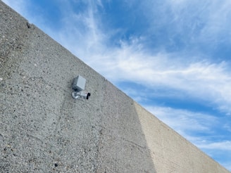 A security camera mounted on a rough, textured concrete wall, angled slightly upwards. The background shows a partly cloudy sky with various shades of blue and white clouds.