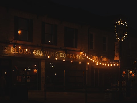 A dimly lit scene of a brewery storefront adorned with strings of warm, glowing lights. The building name, 'Elora Brewing Company', is prominently displayed in illuminated signage. Additional decorations include a lit-up wreath shape on a pole and framed windows.