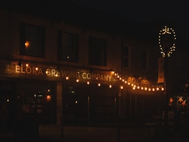 A dimly lit scene of a brewery storefront adorned with strings of warm, glowing lights. The building name, 'Elora Brewing Company', is prominently displayed in illuminated signage. Additional decorations include a lit-up wreath shape on a pole and framed windows.