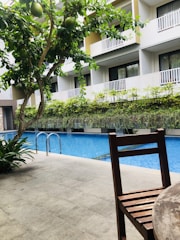 A swimming pool is flanked by a modern building with multiple balconies. There is a presence of green foliage including a tree with large green fruits and various plants, adding to a refreshing atmosphere. A wooden chair is placed in the foreground on a tiled patio.