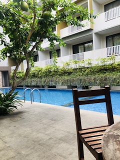 A swimming pool is flanked by a modern building with multiple balconies. There is a presence of green foliage including a tree with large green fruits and various plants, adding to a refreshing atmosphere. A wooden chair is placed in the foreground on a tiled patio.