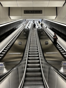 A perspective view of an escalator with metal railings leading upward in a modern indoor setting. Several individuals are visible, ascending and descending, with a prominent sign overhead labeled 'GLACIER'. The lighting is subdued, creating a sleek and industrial atmosphere.