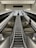 A perspective view of an escalator with metal railings leading upward in a modern indoor setting. Several individuals are visible, ascending and descending, with a prominent sign overhead labeled 'GLACIER'. The lighting is subdued, creating a sleek and industrial atmosphere.