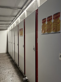 A row of closed white lockers with red borders is neatly arranged along a wall under bright fluorescent lights. The floor is textured with a metallic appearance. There are posters depicting bottles with 'POPPERS' labels attached to the locker doors.
