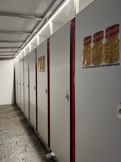 A row of closed white lockers with red borders is neatly arranged along a wall under bright fluorescent lights. The floor is textured with a metallic appearance. There are posters depicting bottles with 'POPPERS' labels attached to the locker doors.