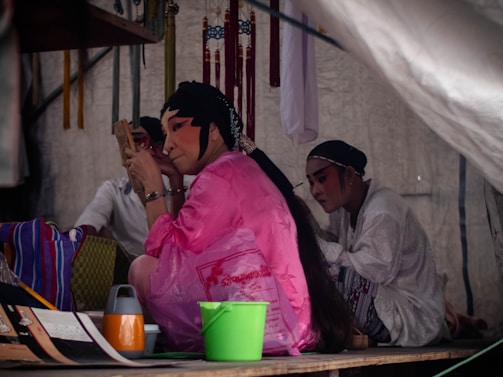 Several individuals are applying traditional facial makeup, likely preparing for a performance. One person in a pink robe is using a handheld mirror while another, dressed in white, watches nearby. Various items such as a green bucket, a striped bag, and some documents are scattered around them. The backdrop includes hanging trinkets and other decorative elements, suggesting a backstage or preparation area.