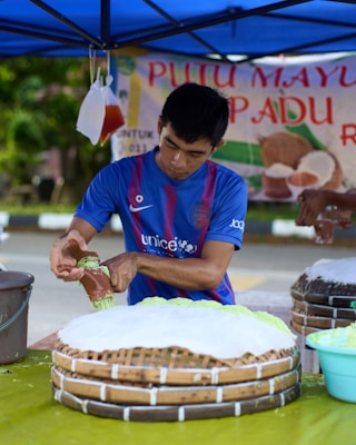 A man wearing a blue sports jersey is preparing traditional food under a blue tent. He seems to be handling green dough or paste and placing it on a large circular woven tray that is partially covered with white powder. There are additional trays and a bucket nearby. Behind him, a sign with the words 'PUTUMAYU' and images of coconuts is partially visible.