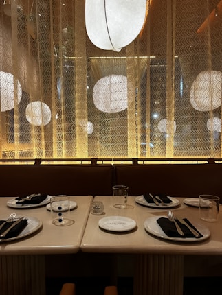 A chic dining area with an elegant and modern table setting. The wooden table is set with plates, cutlery wrapped in black napkins, and glassware for four people. Behind, there is an ambient light setup with large, circular, white light fixtures and decorative panels that enhance the mood.