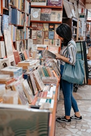 A person is standing in a cozy bookshop, browsing through a book. The books are stacked and arranged on shelves and a table, creating a warm and inviting atmosphere. The person is casually dressed and holding a book while carrying a bag on one shoulder. The bookshop appears to be filled with a variety of books, from novels to art books.