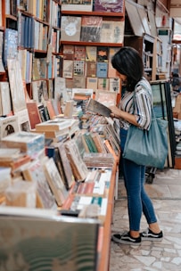 A person is standing in a cozy bookshop, browsing through a book. The books are stacked and arranged on shelves and a table, creating a warm and inviting atmosphere. The person is casually dressed and holding a book while carrying a bag on one shoulder. The bookshop appears to be filled with a variety of books, from novels to art books.