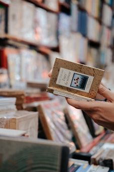 A hand holds a book titled 'Enjoy the Moment' among shelves filled with various books. The focus is on the book in the foreground while the background features blurred stacked books, indicating a library or bookstore setting.