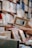A hand holds a book titled 'Enjoy the Moment' among shelves filled with various books. The focus is on the book in the foreground while the background features blurred stacked books, indicating a library or bookstore setting.