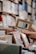 A hand holds a book titled 'Enjoy the Moment' among shelves filled with various books. The focus is on the book in the foreground while the background features blurred stacked books, indicating a library or bookstore setting.