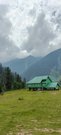 A large, green-roofed house stands in the foreground, surrounded by a vast meadow. The lush greenery extends into dense forested hills, with towering pine trees and cascading slopes. In the background, mountain peaks are partially obscured by a thick layer of dramatic, overcast clouds, creating a contrast between the dark and light areas.