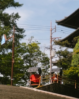 Two rickshaws with red covers are parked on a raised platform near a traditional-style building with ornate roofing. Lush green trees surround the area, and a utility pole with wires is visible in the background. A pedestrian crossing sign is affixed to a tall pole.