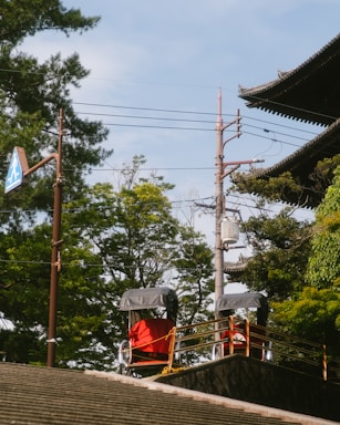 Two rickshaws with red covers are parked on a raised platform near a traditional-style building with ornate roofing. Lush green trees surround the area, and a utility pole with wires is visible in the background. A pedestrian crossing sign is affixed to a tall pole.