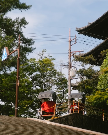 Two rickshaws with red covers are parked on a raised platform near a traditional-style building with ornate roofing. Lush green trees surround the area, and a utility pole with wires is visible in the background. A pedestrian crossing sign is affixed to a tall pole.