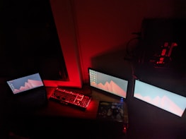 A setup featuring three monitors and a keyboard emitting a red glow, creating a moody atmosphere. The monitors display a minimalistic wallpaper with red geometric shapes, and the room is dimly lit with red lighting. Cables are visible around the desk, adding to the tech-heavy feel.