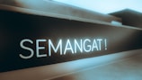 A close-up of a sign or board with the word 'SEMANGAT!' written in bold, bright letters, which appears to be illuminated or glowing against a dark background. The setting conveys a sense of motivation or encouragement.