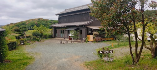 A rustic building surrounded by greenery in a rural setting. The building has a traditional design with a dark wooden exterior, large windows, and a sloped roof. In front of the building, there are outdoor seating arrangements, plants, and a visible 'OPEN' sign. The surrounding area includes gravel pathways, grass, trees, and distant hills under a cloudy sky.