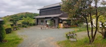 A rustic building surrounded by greenery in a rural setting. The building has a traditional design with a dark wooden exterior, large windows, and a sloped roof. In front of the building, there are outdoor seating arrangements, plants, and a visible 'OPEN' sign. The surrounding area includes gravel pathways, grass, trees, and distant hills under a cloudy sky.
